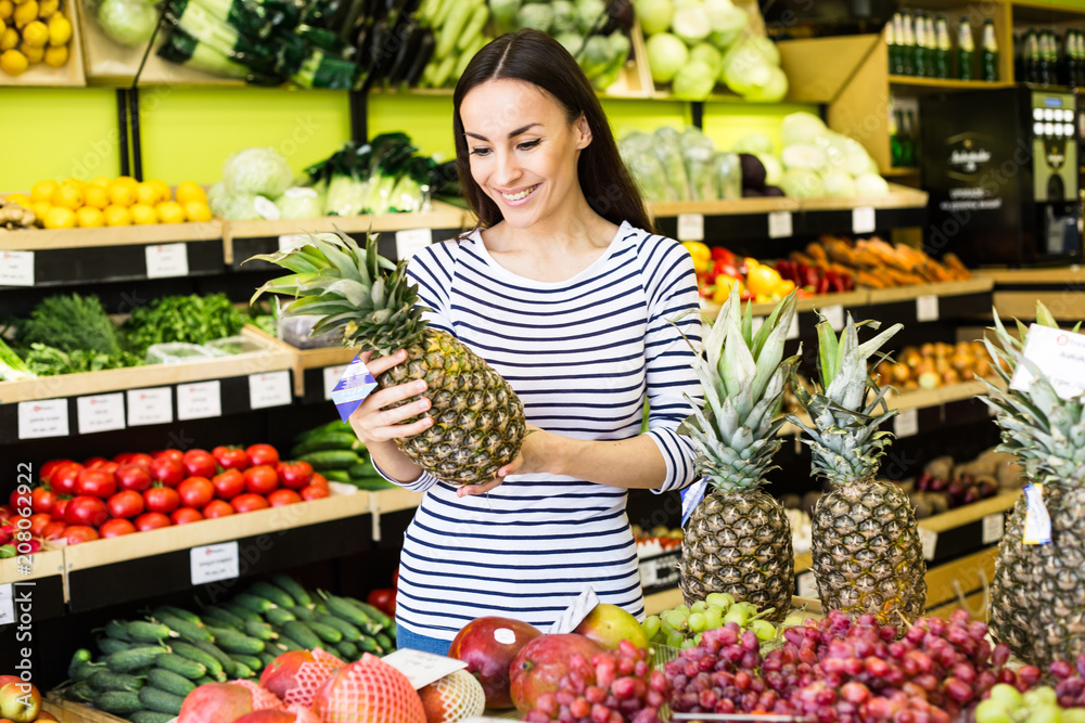 Attractive smiling young girl in casual clothes selects fruits and vegetables from a shelf in a grocery store.