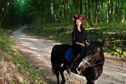 Mystical girl in wreath wear in black at horse in wood.