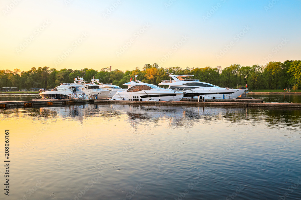 Fototapeta premium Luxury yachts docked in marina at colorful sunset. Marine parking, modern motor boats moored in water bay at pier. Summer view of river bank, embankment and superyachts in sunshine. Sunny evening.