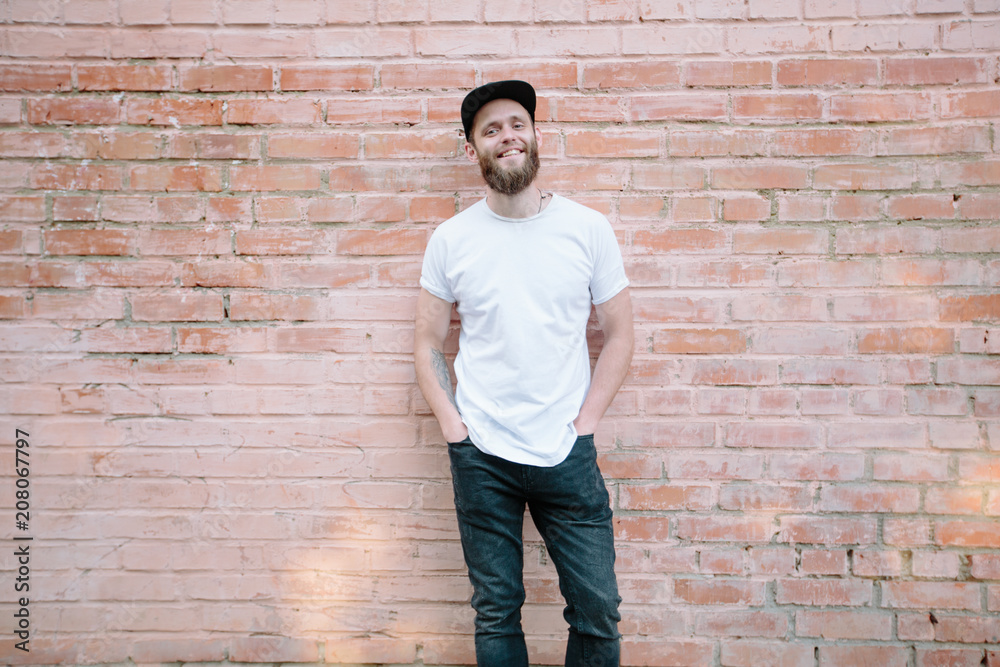 Hipster handsome male model with beard wearing white blank t-shirt and a baseball cap with space for your logo or design in casual urban style