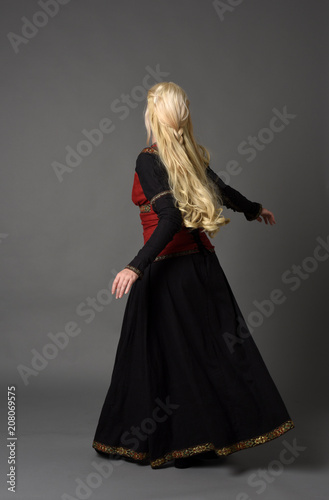full length portrait of pretty blonde lady wearing  a red and black fantasy medieval gown. standing pose on grey background.