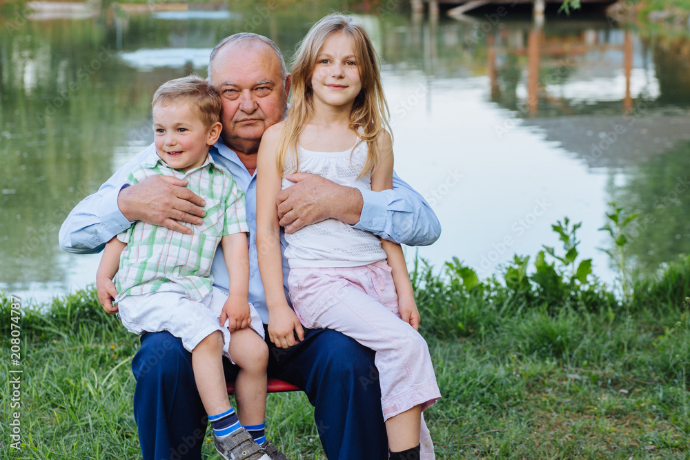 Fototapeta premium Sweet family portrait of three happy relatives - grandfather hugging with his granddaughter and grandson over lake background at summer time.