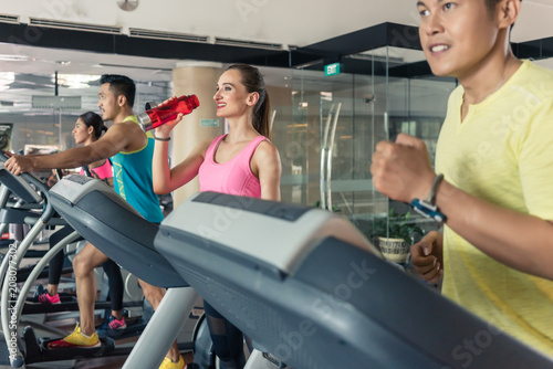 Cheerful beautiful woman with a healthy lifestyle drinking water during training on treadmill in a trendy fitness club