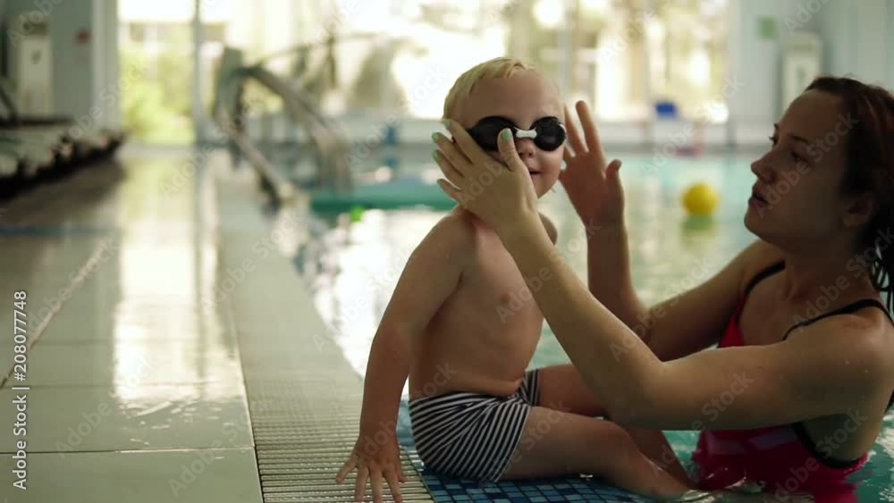 A small swimmer prepares to swim. The boy sits on the edge of the pool, his mother puts on his glasses for swimming. Waving his feet on the water. Indoors A small swimmer prepares to swim. The boy sits on the edge of the pool, his mother puts on his glasses for swimming. Waving his feet on the water. Indoors