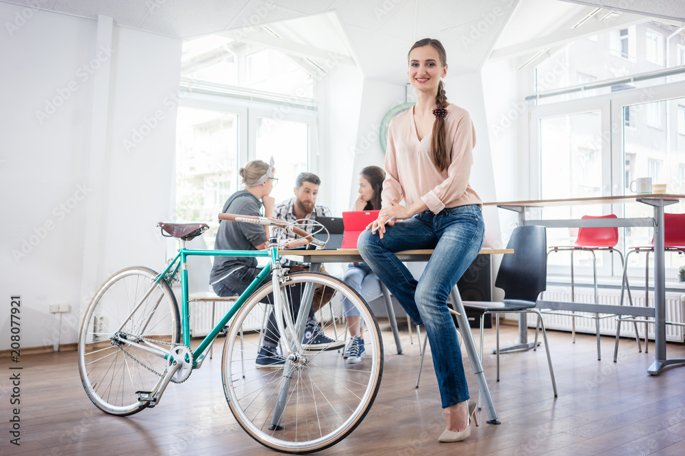 Full length of a beautiful and confident young woman wearing blue jeans, while sitting on a desk near her commuter bike in a co-working office space Full length of a beautiful and confident young woman wearing blue jeans, while sitting on a desk near her commuter bike in a co-working office space