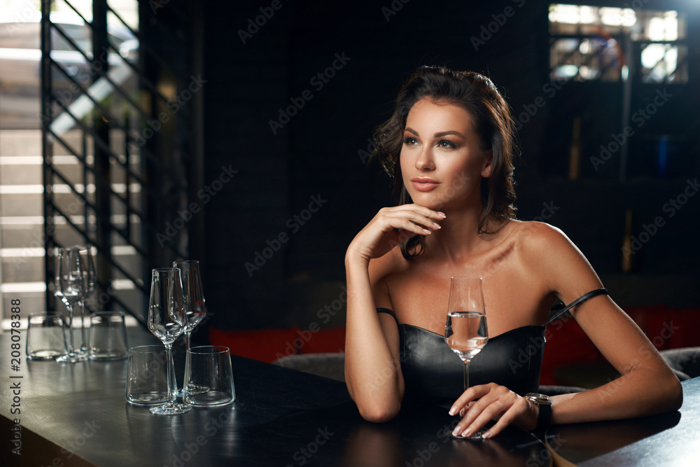 Elegant young woman sitting in bar and having good time alone. Pretty brunette girl in black leather dress. Lady drinking glass of wine. Elegant young woman sitting in bar and having good time alone. Pretty brunette girl in black leather dress. Lady drinking glass of wine.