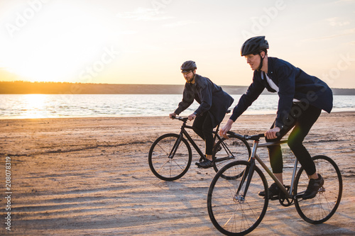 Two young men riding bicycles on the beach on the background of an orange sunsetting sky