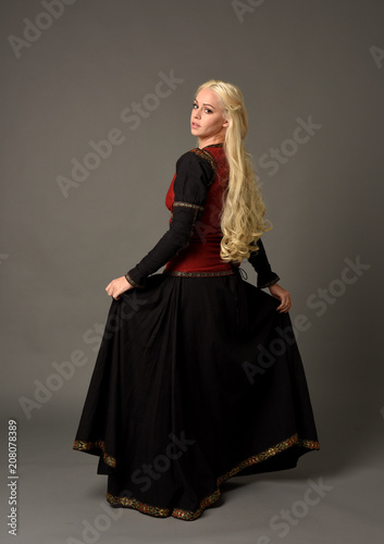full length portrait of pretty blonde lady wearing  a red and black fantasy medieval gown. standing pose on grey background.