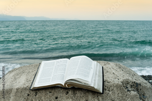 Bible open on top of a large stone in front of the green sea and an orange sky. Copy space. photo