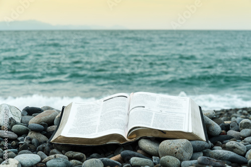 Fotomurale Bible open on top of small stones in front of the green sea and an orange sky