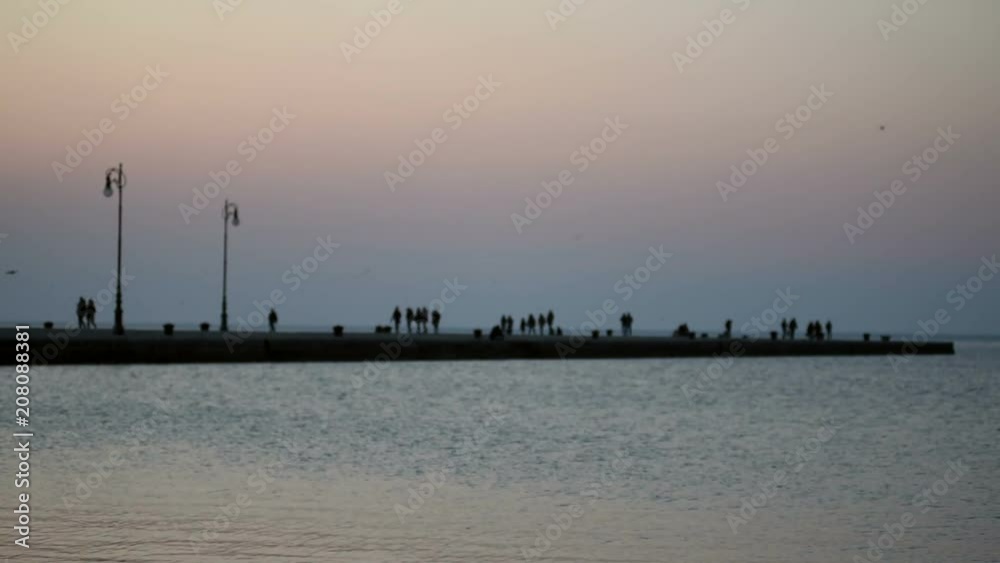 Trieste, Italy - Intentionally blurred view of people strolling on the Molo Audace pier at sunset. Small waves of the sea on the foreground, silhouetted pier with people slowly walking and streetlamps Trieste, Italy - Intentionally blurred view of people strolling on the Molo Audace pier at sunset. Small waves of the sea on the foreground, silhouetted pier with people slowly walking and streetlamps