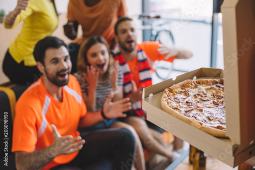 close up shot of pizza in box and excited group of friend sitting on sofa at home photo