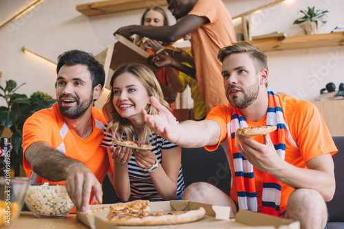 young man pointing by hand and his friends eating pizza and watching soccer match at home photo