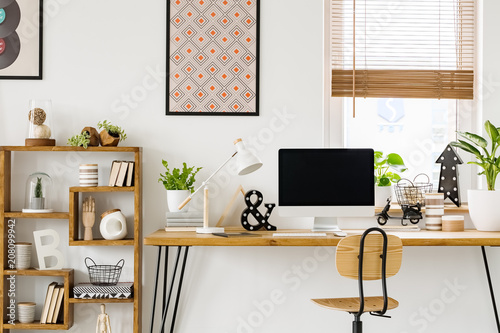 Real photo of a desk with a computer screen, lamp and ornaments standing with a chair next to a shelf with more ornaments in a work room with posters on a wall and window with blinds