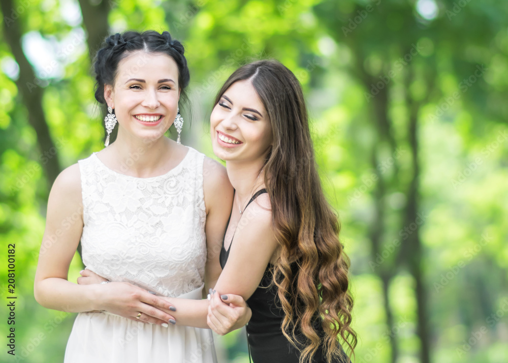 Portrait of two young beautiful women laughing and hugging each other in green summer park. Pretty females bride and bridesmaid smiling and looking into camera. Copy spece Portrait of two young beautiful women laughing and hugging each other in green summer park. Pretty females bride and bridesmaid smiling and looking into camera. Copy spece