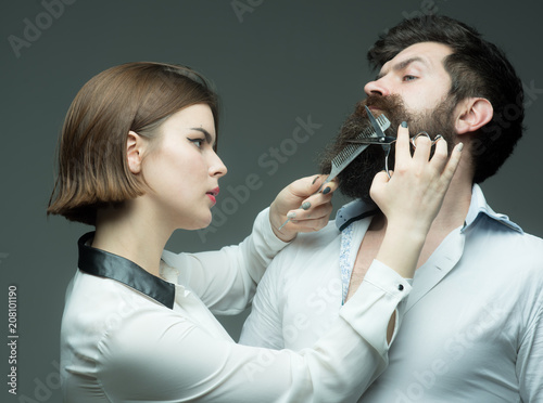 Barbershop or hairdresser concept. Woman hairdresser cuts beard with scissors. Guy with modern hairstyle visiting hairdresser. Man with long beard, mustache and stylish hair, grey background