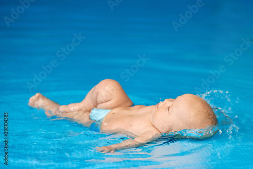 Little baby boy lying and splashing in blue swimming pool in water.