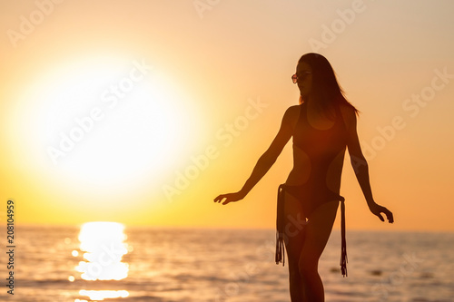 Pretty young woman with long dark hair near the ocean tanning in black swimwear. concept of happy holiday and resort time. silhouette over sunset