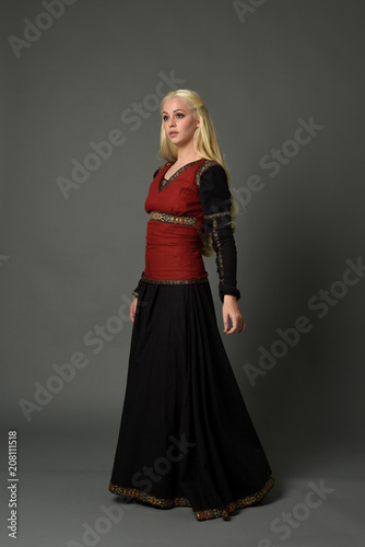 full length portrait of pretty blonde lady wearing  a red and black fantasy medieval gown. standing pose on grey background.