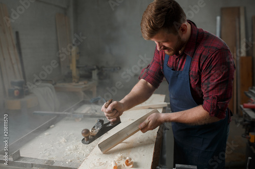 Master carpenter in shirt and apron strokes plane in workshop. Close up