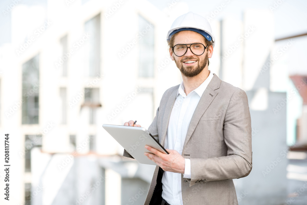 Handsome engineer working with tablet standing on the structure site with a new house on the background Handsome engineer working with tablet standing on the structure site with a new house on the background