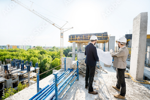 Two engeneers or architects working with house drawings standing together on the structure during the construction process outdoors