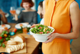 Crop close-up young woman standing at home and holding white bowl with green salad while eating with friends