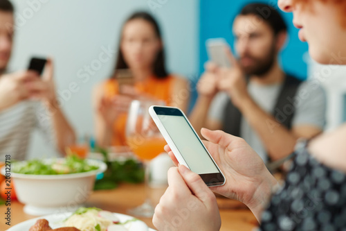 Crop close-up female using smartphone while sitting at table and eating together with friends