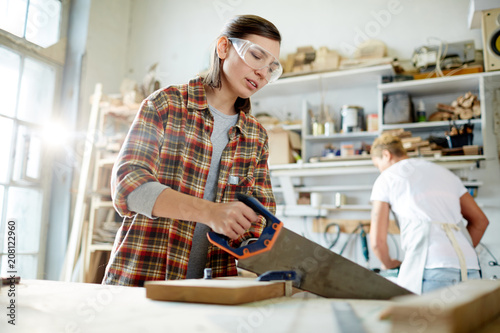 Adult woman in safety goggles using saw to cut timber board while working in carpenter workshop