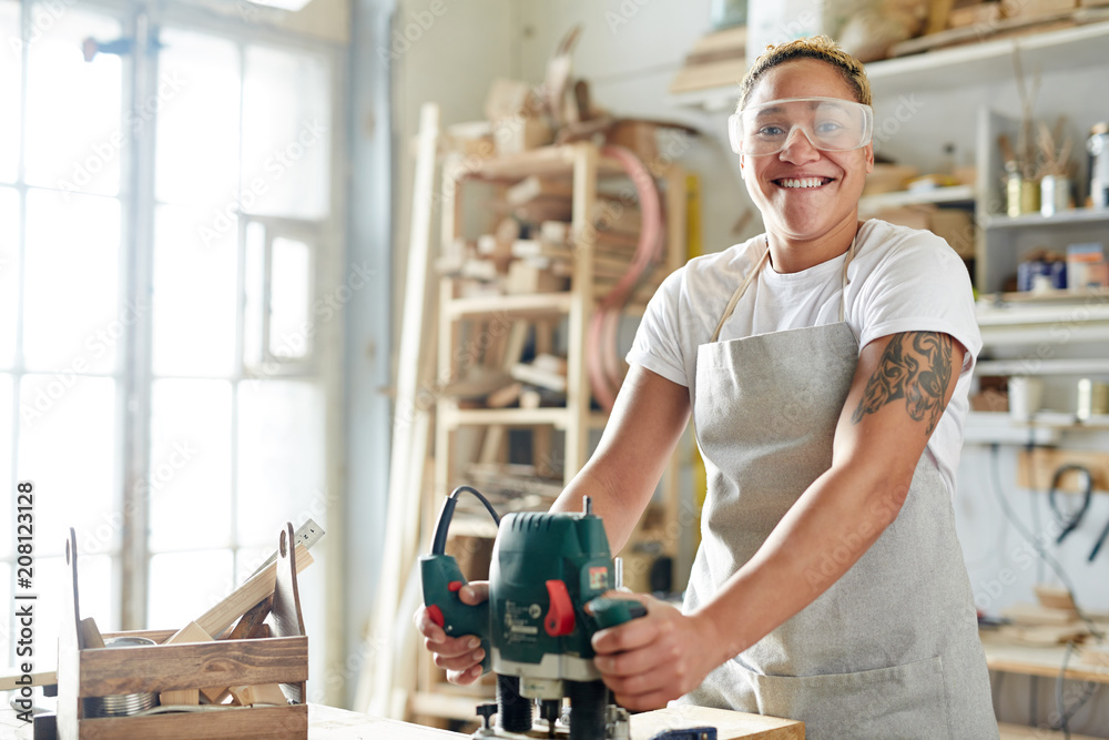 Female carpenter in apron and protective glasses smiling and looking at camera while using electric saw in workshop. 