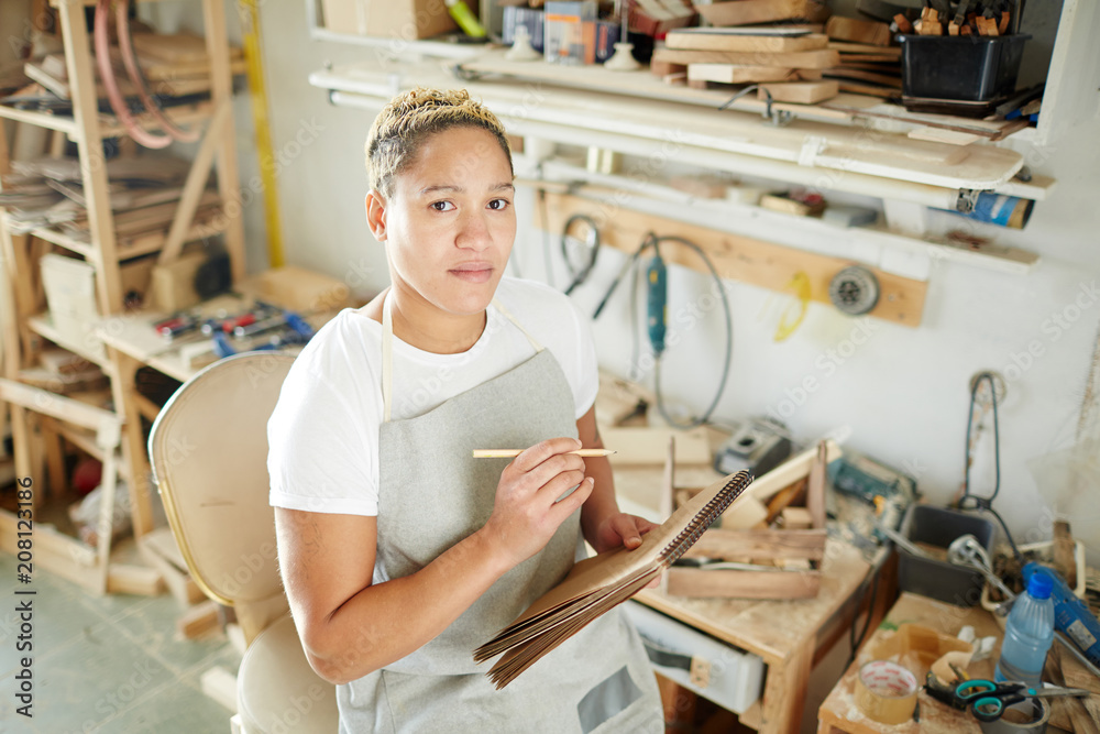 Woman in apron making notes in notebook and looking at camera while working in carpenter workshop. Woman in apron making notes in notebook and looking at camera while working in carpenter workshop.