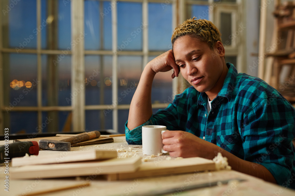 Tired female woodworker holding mug with aromatic hot beverage while sitting near workbench in workshop. Tired female woodworker holding mug with aromatic hot beverage while sitting near workbench in workshop.