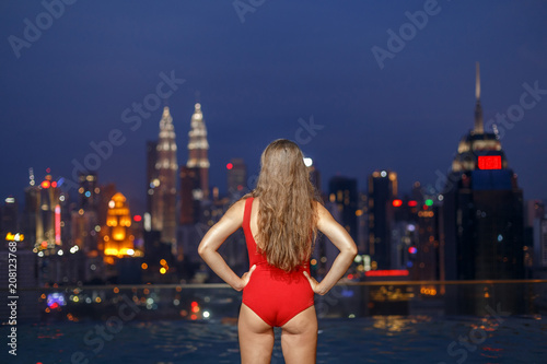 Beautiful woman portrait in red swimsuit, in swimming rooftop pool, at night city landscape of Kuala Lumpur, Malaysia