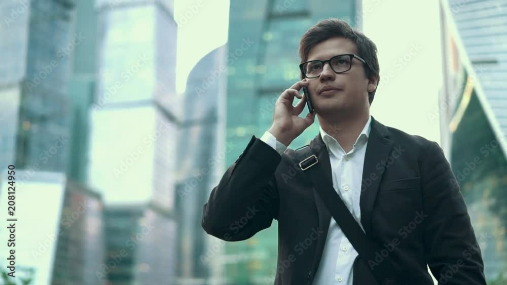 Handsome young businessman in a dark suit and glasses talking calmly on his smartphone on a cloudy summer day in Moscow city. Slider medium shot Handsome young businessman in a dark suit and glasses talking calmly on his smartphone on a cloudy summer day in Moscow city. Slider medium shot