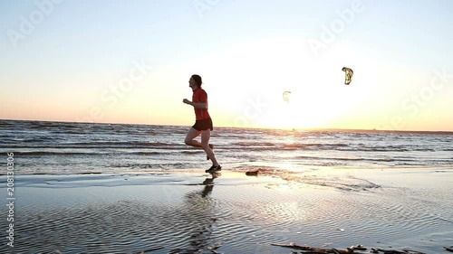 Cheerful male runner jumping in the water against the sunset and kitesurfers, slow motion