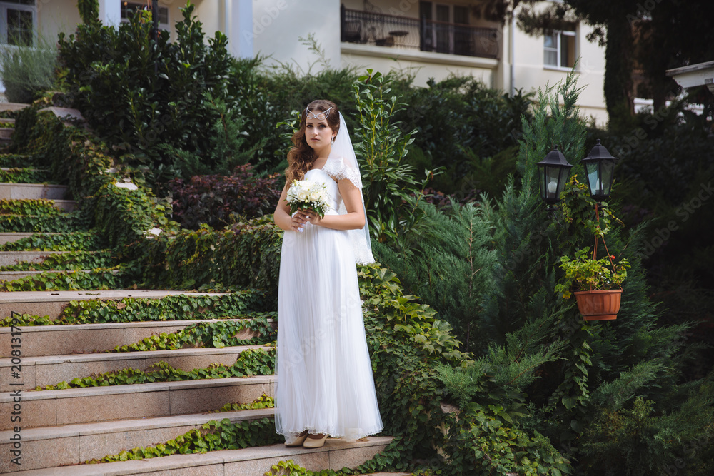 The beautiful young bride stands on stubble stairs in a dress of Greek style, her hair is curly, decorated with beads, she is holding a wedding bouquet of white roses.