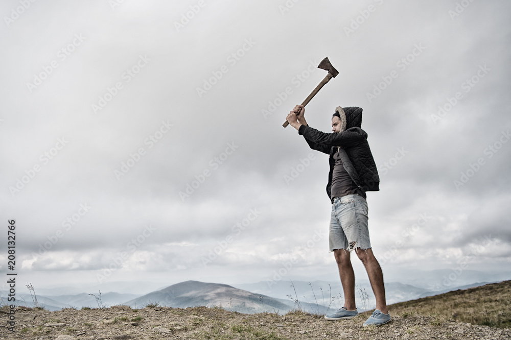 Hipster with beard holds axe while stand on top of mountain, sky on background, copy space. Lumberjack brutal and bearded raising axe. Man in hat and jacket conquered top. Brutal lumberjack concept