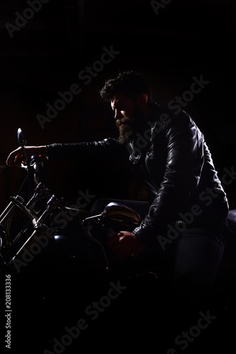 Night racer concept. Macho, brutal biker in leather jacket riding motorcycle at night time, copy space. Man with beard, biker in leather jacket sitting on motor bike in darkness, black background.