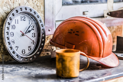 Lunch time at the production plant or a construction site for people who are working. 
Still life clock showing the time lunch, an orange helmet, a dirty orange plastic Cup, close-up