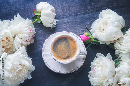 A cup of coffee and white peonies on dark wooden background. Top view, copy space. Toned image