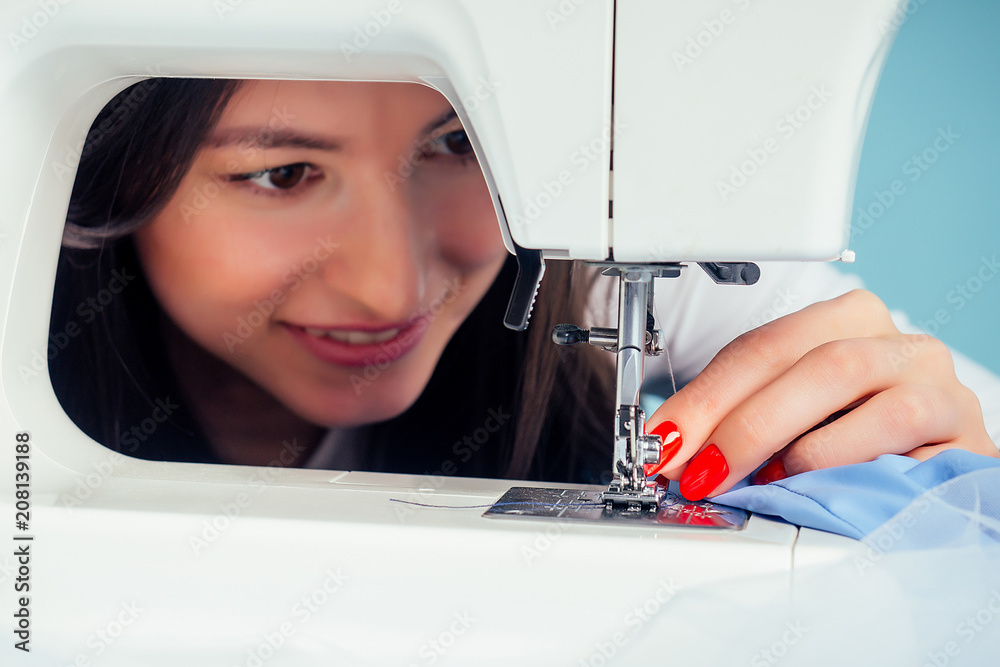 close-up portrait attractive woman seamstress tailor ( dressmaker) thread the needle on the sewing machine on a blue background in the studio. The concept of creating a new collection of clothes