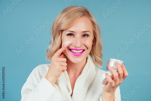 attractive and smiling blonde female person in a cotton white bathrobe applies moisturizer cream on face in the studio on a blue background . concept of skin care and spa