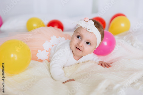 The baby is lying on the bed and looking away with interest and a smile. The girl is dressed in a beautiful dress and crawls on a white bed.