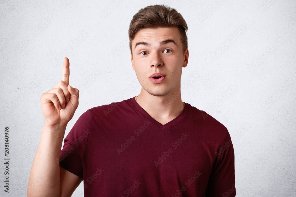 Stunned male teenager with suprised expression keeps fore finger raised, dressed in casual t shirt, has trendy hairstyle, poses against concrete white wall. Amazed guy indicates upwards. Advertisement Stunned male teenager with suprised expression keeps fore finger raised, dressed in casual t shirt, has trendy hairstyle, poses against concrete white wall. Amazed guy indicates upwards. Advertisement