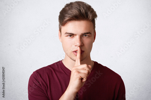 Headshot of handsome hipster guy looks secretly at camera, keeps fore finger on lips, demonstrates silence sign, dressed casually, isolated over white background. People and conspiracy concept