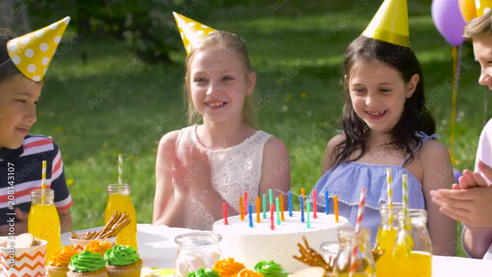 holidays, childhood and celebration concept - happy kids blowing out candles on birthday cake sitting at table at summer garden party