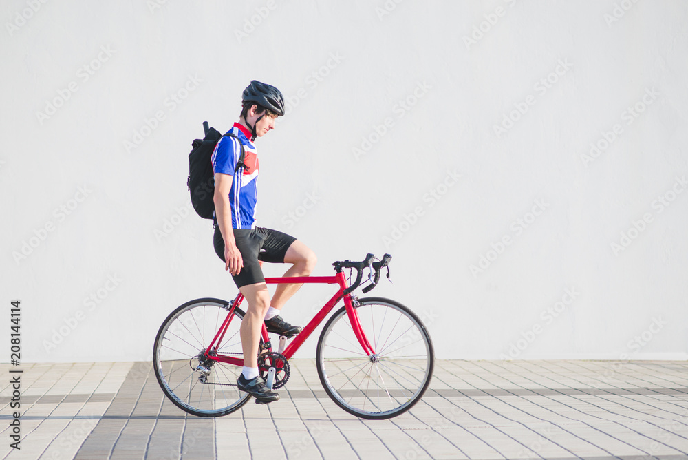 Fototapeta premium Athlete cyclist in cycling clothes and a helmet riding a bike without hands on the background of a light wall. Sports concept. Man with a bicycle on the flush wall.