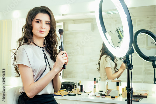 charming beautiful woman make-up artist (visagiste ) sitting in front of a mirror and holding a brushes cosmetics make-up on face in studio