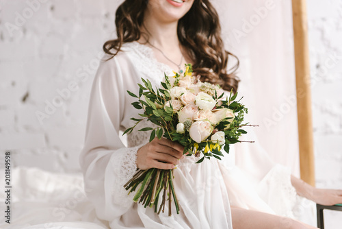 Portrait of a young charming bride in front of a wedding ceremony with a big beautiful bouquet in white lingerie. on a large bed. Close-up on a bouquet