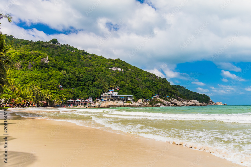 Fototapeta premium Tropical sandy beach with palms, houses and woodean boats in the background and blue ocean in the foreground during a bright sunny day, Haad Rin Beach, Koh Phangan, Thailand